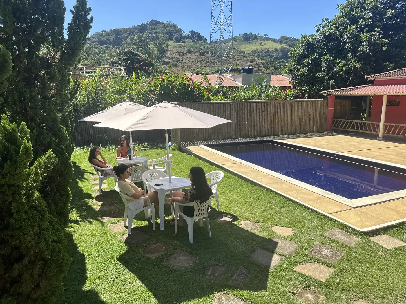 Amigas sob guarda-sóis no gramado ao lado da piscina, com morros ao fundo