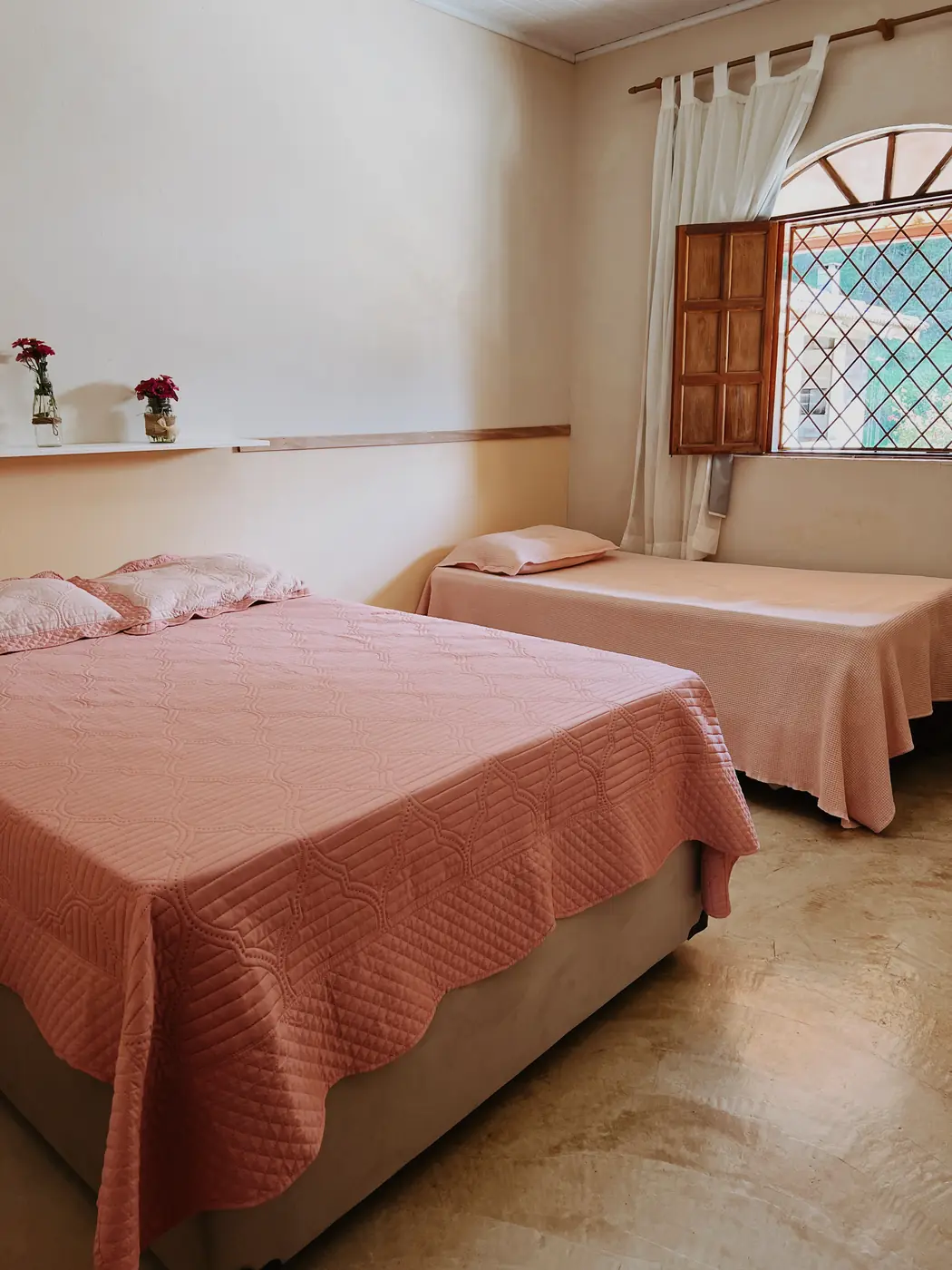 Quarto com cama de casal e de solteiro, colchas rosadas e janela de madeira